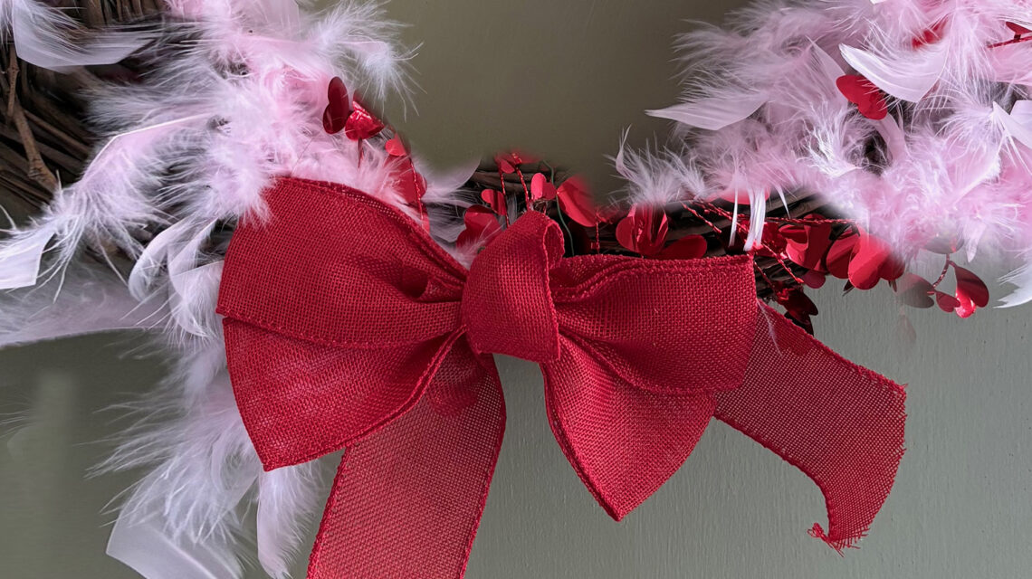 Tight crop on the ribbon of a wreath on a gray-green door. The wreath is a brown wicker circle with pale pink maribou feathers and a red metallic heart garland wound around it. There's a red ribbon bow at the bottom.